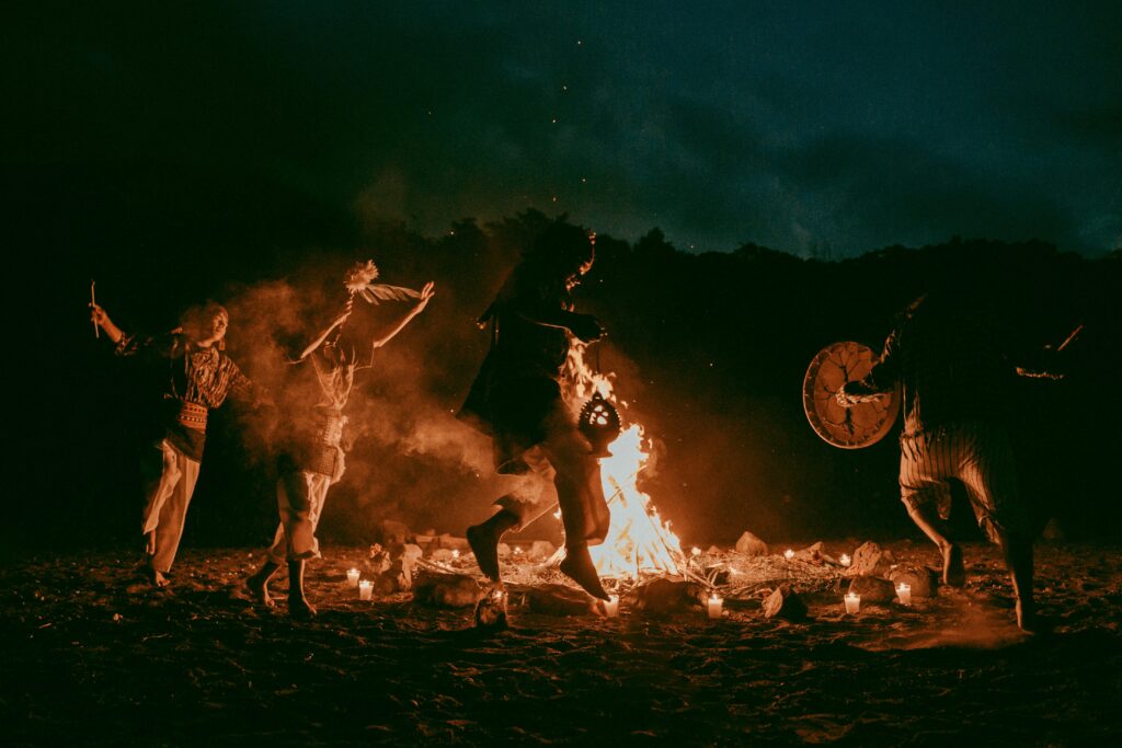Four people are dancing in the night around a lit bonfire, running in circle around it. They're wearing traditional outfits and accessories, and there are lit candles and rocks all around the fire 